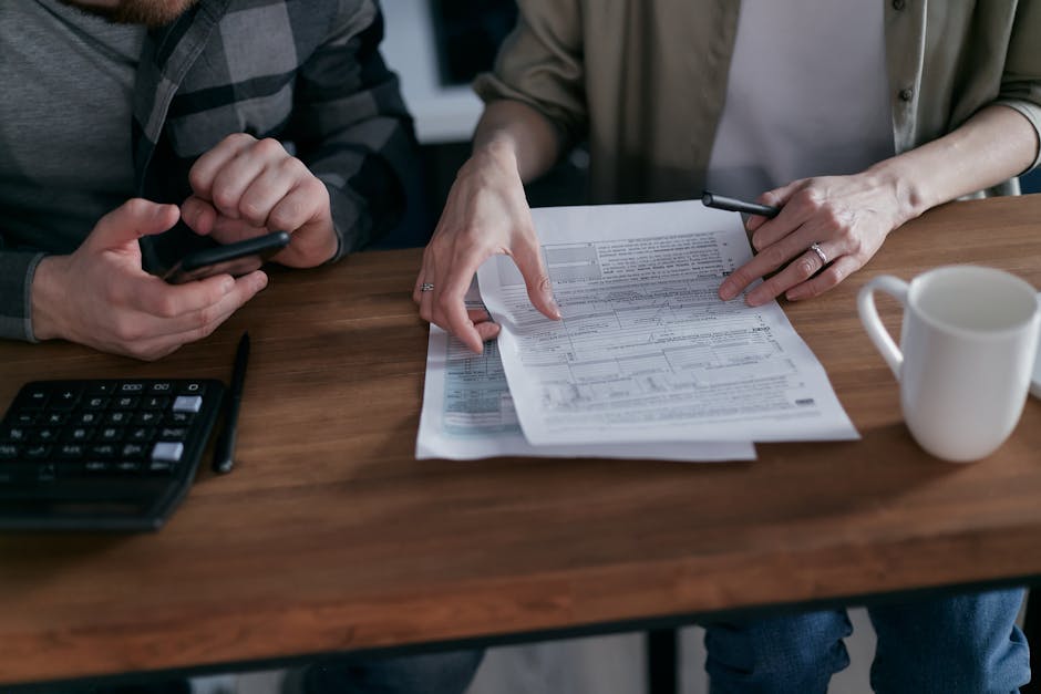 Two adults reviewing financial documents, using a calculator and smartphone at a wooden table.